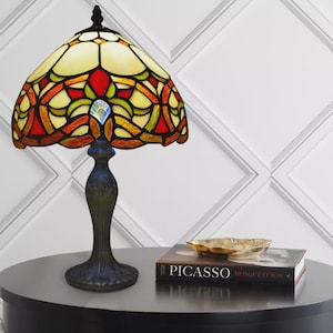 May include: A stained glass table lamp with a colorful shade featuring red, yellow, and green hues. The lamp has a dark blue, ornate base and sits on a dark round table. A book titled "Picasso" and a gold-colored bowl are also on the table.