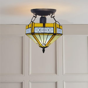 May include: A yellow and white stained glass ceiling light fixture. The fixture has a geometric design with black metal accents and a decorative finial. The light is suspended from the ceiling by a chain.