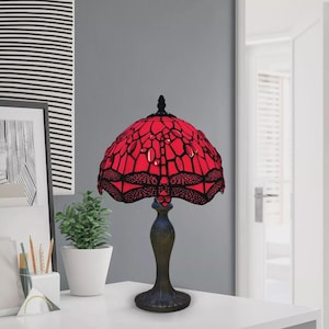 May include: A red and black stained glass table lamp with a dragonfly design. The lamp has a bronze-coloured base and a red shade with black accents. The lamp is on a white surface.