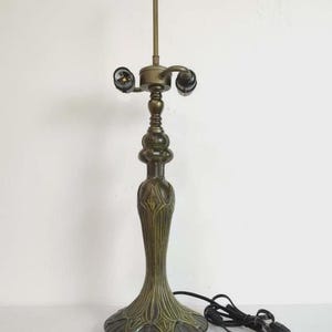 May include: A vintage-style table lamp with a bronze finish. The lamp features a detailed base, a decorative central column, and a two-bulb socket. The lamp is connected to a black power cord.