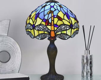 May include: A stained glass table lamp with a dragonfly design. The lamp features a blue, yellow, and purple stained glass shade. The base is dark brown with an ornate design. The lamp is on a glass surface.