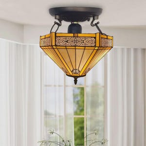 Può includere: Un lampadario decorativo con paralume in vetro colorato. Il paralume è di colore giallo caldo con motivi geometrici e dettagli in metallo marrone scuro. L'apparecchio è appeso a un supporto a soffitto marrone scuro.