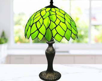 May include: A decorative table lamp with a green stained glass shade in a leaf pattern. The base is dark bronze with an ornate design. The lamp is on a white surface, with a blurred background of a window and a kitchen.
