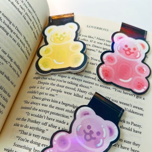 Pu&ograve; includere: Tre segnalibri a forma di orso gommoso di diversi colori e design. I segnalibri sono gialli, rosa e viola. I segnalibri sono sopra un libro con testo su Harry Potter.