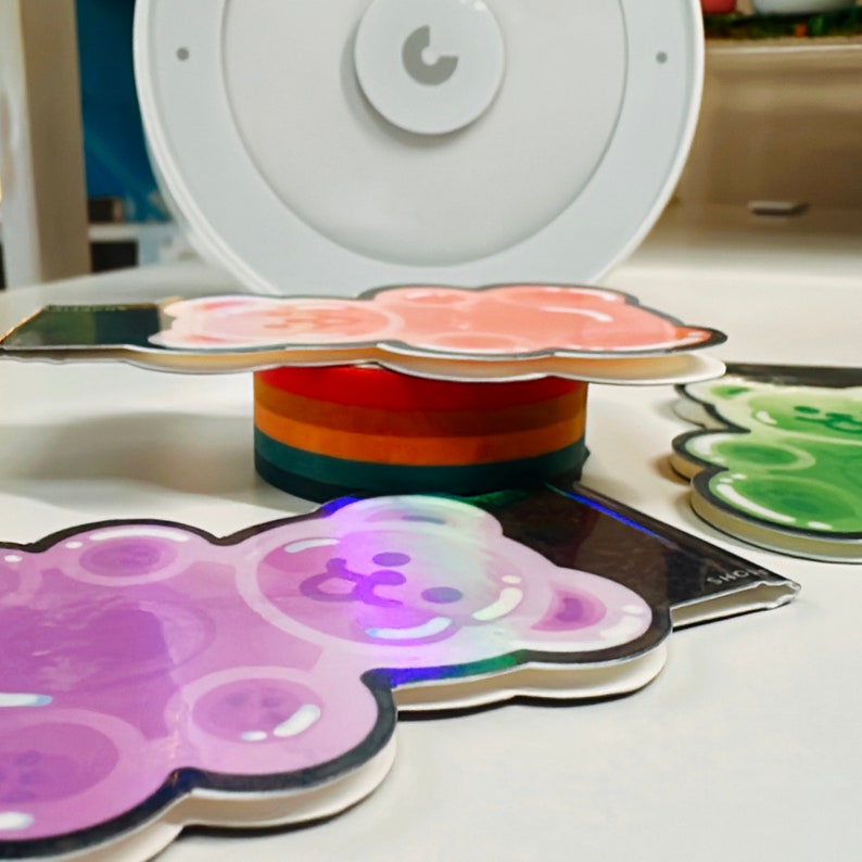 Pu&ograve; includere: Quattro adesivi a forma di orso gommoso colorati, uno rosa, uno viola, uno verde e uno arcobaleno, sovrapposti. Gli adesivi sono su una superficie bianca.