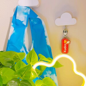 Peut inclure: Un sac fourre-tout bleu et blanc &agrave; motif de nuages est suspendu &agrave; un crochet blanc en forme de nuage. Un porte-cl&eacute;s Kudos rouge et jaune est suspendu &agrave; un autre crochet nuage. Une lumi&egrave;re n&eacute;on en forme de nuage et une plante verte sont au premier plan.