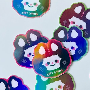 May include: A set of five iridescent holographic stickers featuring a cartoon character with a black and white face and a rainbow-colored background. The stickers have the text "# KEEP GOING #" on them.