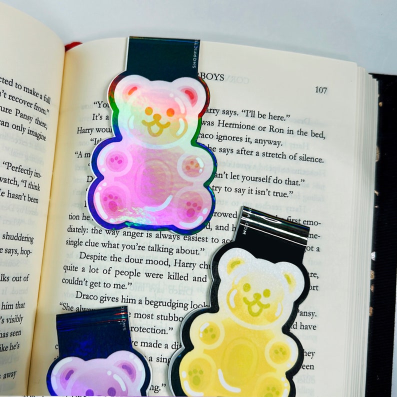 Pu&ograve; includere: Due segnalibri a forma di orso gommoso olografici. Uno &egrave; rosa con un contorno blu e l'altro &egrave; giallo con un contorno nero. Entrambi hanno un simpatico viso di orso da cartone animato.