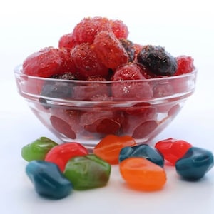 Puede incluir: Un bol de vidrio lleno de caramelos gomosos rojos y negros. El bol está sobre una superficie blanca. Hay varios caramelos gomosos de colores esparcidos alrededor del bol.