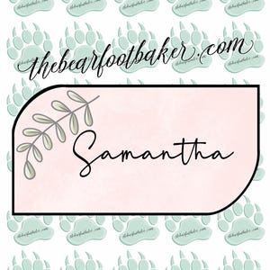 May include: A light pink rectangular sticker with the name "Samantha" in cursive, framed by a black border and a sprig of leaves. The background features repeating light blue paw prints and the text "thebearfootbaker.com".