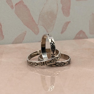 May include: Three silver rings stacked together, each featuring a repeating engraved floral pattern around the band. The metal has a polished finish with darkened recessed areas that highlight the intricate flower and leaf design details.