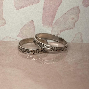 May include: Two silver-toned metal rings featuring a repeating embossed floral pattern with small petals and vertical lines. The rings have a polished finish and are positioned so that one ring partially overlaps the other, showing the band design.