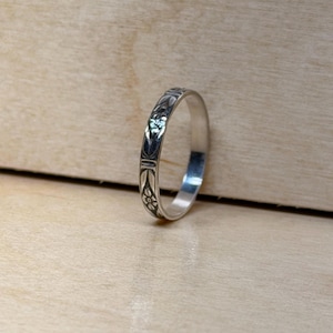 May include: A silver ring with an engraved floral pattern. The ring is circular and has a detailed design that wraps around the band. The ring is set against a light brown wooden surface.