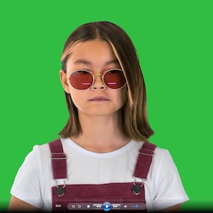 May include: A person wearing round, gold-framed sunglasses with red lenses. They are wearing a white t-shirt and burgundy dungarees against a bright green backdrop. The image is a portrait.