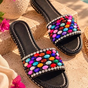 May include: A pair of black slide sandals featuring a wide strap adorned with rows of multicolored oval beads and clear rhinestones along the edges. The footbed is black with a stitched border, and the sole is detailed with small silver studs.