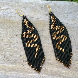 May include: A pair of long, black and gold beaded earrings with a snake design. The earrings are made with small, round beads and have a tassel-like fringe at the bottom.