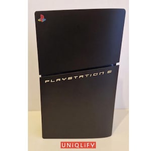 May include: A matte black gaming console featuring a diagonal split design. The top section displays a colorful PlayStation logo, while the lower section features the gold text PLAYSTATION 5. A red rectangular logo with the text UNIQLIFY is visible.