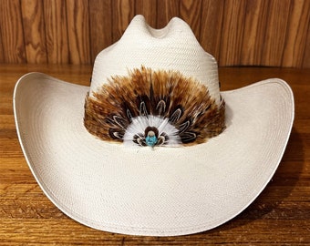 Puede incluir: Un sombrero de vaquero blanco con un adorno de plumas marrones y blancas y un acento de piedra turquesa.