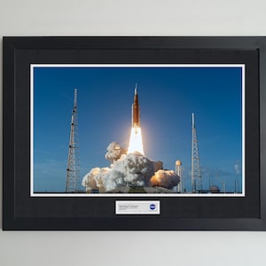 May include: Framed print of a rocket launching into a clear blue sky, with billowing white smoke and flames. The rocket is orange and white. Two tall towers flank the rocket. The print is in a black frame.