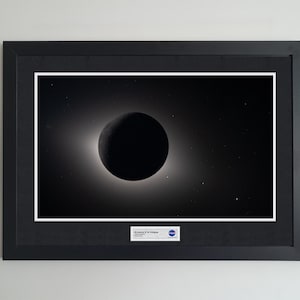 May include: A framed black and white photograph of a solar eclipse featuring a dark circular silhouette surrounded by a glowing halo of light. A small white rectangular label at the bottom includes the NASA logo and the text Artemis II in Eclipse.
