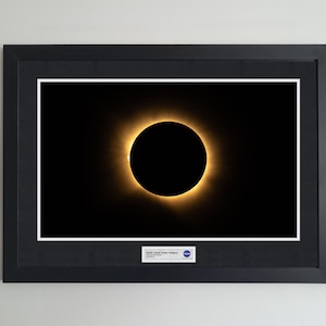 Könnte beinhalten: Ein gerahmtes Foto einer totalen Sonnenfinsternis mit einem dunklen zentralen Mond, der von einer leuchtend goldenen Korona umgeben ist. Das Bild befindet sich in einem schwarzen Rahmen mit weißem Passepartout und einem Etikett 2017 Total Solar Eclipse.