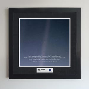 Op de afbeelding: Ingelijste print met een foto van een lichtblauwe stip in de ruimte, met een donkerblauwe en witte achtergrond. De print bevat de quote "Kijk nog eens naar die stip. Dat is hier. Dat is thuis. Dat zijn wij." en de titel "Pale Blue Dot".