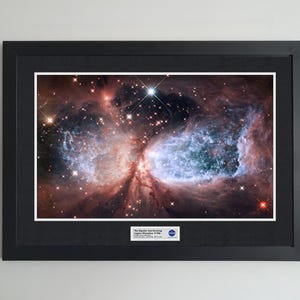 May include: Framed print of a nebula, showcasing vibrant colors of red, blue, and white. The image is set within a black frame with a white mat. A small plaque with text and the NASA logo is visible at the bottom.