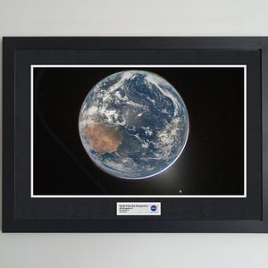 May include: A framed print depicting Earth from space, with blue oceans, brown landmasses, and white clouds against a black backdrop. The print is in a black frame with a white mount, and includes a small text label.