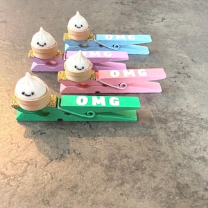 May include: A collection of decorative clothespins in various pastel colors, each adorned with a miniature dumpling figurine. The word "OMG" is written on each pin in white. The dumplings have smiling faces.