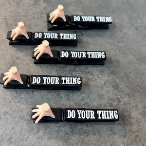 May include: Black clothespins with the text "DO YOUR THING" in white. Each pin has a small, light-colored hand attached. The clothespins are arranged on a gray surface.