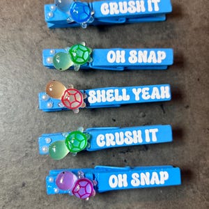 May include: Five light blue wooden clothespins with white text and turtle embellishments. The text on the clothespins reads "CRUSH IT", "OH SNAP", and "SHELL YEAH". Each pin has a different colored turtle and small pearl accents.