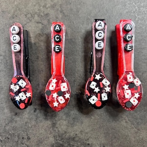 May include: Four red and black spoon-shaped hair clips. Each clip has the word "ACE" in white letters. The spoon bowls are filled with playing card and star-shaped glitter. The clips are on a gray surface.