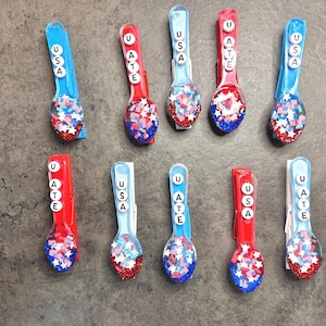 May include: A collection of patriotic-themed spoons. Each spoon is decorated with red, white, and blue glitter and star-shaped confetti. The handles feature the letters "USA" in white. The spoons are arranged on a gray surface.