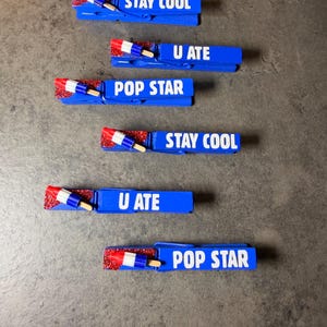 May include: Blue clothespins with white text that reads "STAY COOL", "U ATE", and "POP STAR". Each pin has a red glitter accent and a miniature popsicle. Decorative and fun.