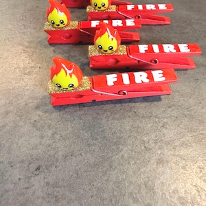May include: Red wooden clothespins with the word "FIRE" in white lettering. Each pin features a cartoon flame with a smiling face and gold glitter accents. The flames are red and yellow.