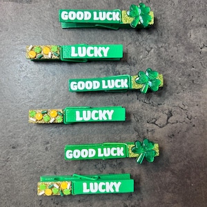 May include: Green wooden clothespins with the words "GOOD LUCK" and "LUCKY" in white. Some pins have a shamrock and glitter embellishments. The clothespins are arranged on a gray surface.