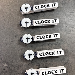 May include: Five white wooden clothespins with a clock face design and the words "CLOCK IT" in black. The clock faces are white with black hands, and the clothespins have a dark blue glitter accent.