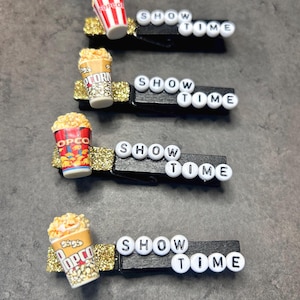 May include: Four black wooden clothespins with gold glitter accents, each topped with a miniature popcorn bucket and the words "SHOW TIME" spelled out in white letter beads. The popcorn buckets have different designs.