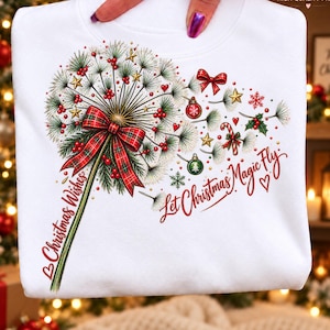 May include: A white sweatshirt featuring a graphic of a dandelion made of pine needles, red berries, and gold stars. A red plaid bow is tied to the stem. The text reads "Christmas Wishes" and "Let Christmas Magic Fly" in red script.