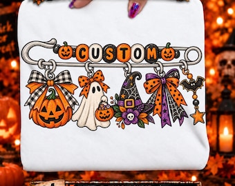 May include: A white t-shirt featuring a graphic of a large silver safety pin with the word CUSTOM spelled out in orange and black beads. Hanging from the pin are charms including a jack-o'-lantern, a ghost, a witch hat, and a bat.