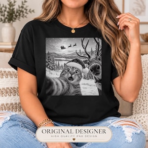 May include: A black t-shirt featuring a black and white graphic print of a cat wearing a Santa hat taking a selfie with a reindeer in a snowy forest landscape, with a silhouette of Santa Claus and his sleigh flying in the background sky.