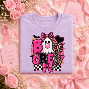 May include: A light purple t-shirt featuring a graphic of a white ghost with a pink nose, wearing a leopard print bow. The text reads BOO CREW in pink and leopard print letters, accented with black bats, spiderwebs, and a checkered pattern.