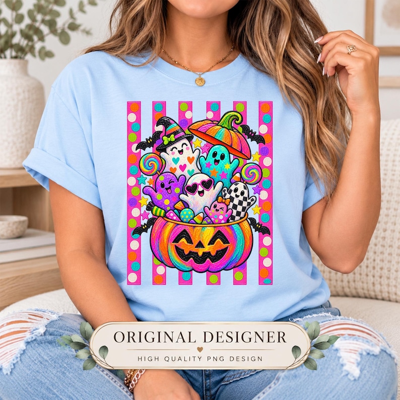May include: A light blue t-shirt featuring a colorful graphic of a large jack-o'-lantern filled with various cartoon ghosts, candy, and lollipops. The design includes a background of vertical pink stripes with colorful polka dots and small bats.