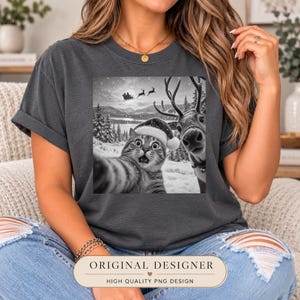 May include: A charcoal gray t-shirt featuring a black and white graphic print of a cat wearing a Santa hat taking a selfie with a reindeer. The background of the print shows a snowy winter landscape with a silhouette of Santa's sleigh in the sky.