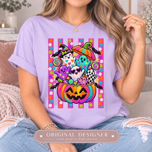 May include: A light purple t-shirt featuring a colorful graphic of a large jack-o'-lantern filled with various cartoon ghosts, bats, lollipops, and candy. The design is set against a background of vertical pink stripes with colorful polka dots.