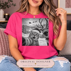 May include: A bright pink short-sleeved t-shirt featuring a black and white graphic print of a cat wearing a Santa hat taking a selfie with a reindeer in a snowy landscape with Santa's sleigh flying in the background sky.