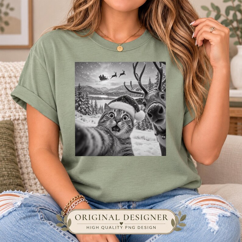 May include: A sage green t-shirt featuring a black and white graphic print of a cat wearing a Santa hat taking a selfie with a reindeer in a snowy landscape, with Santa's sleigh flying in the background under a starry night sky.