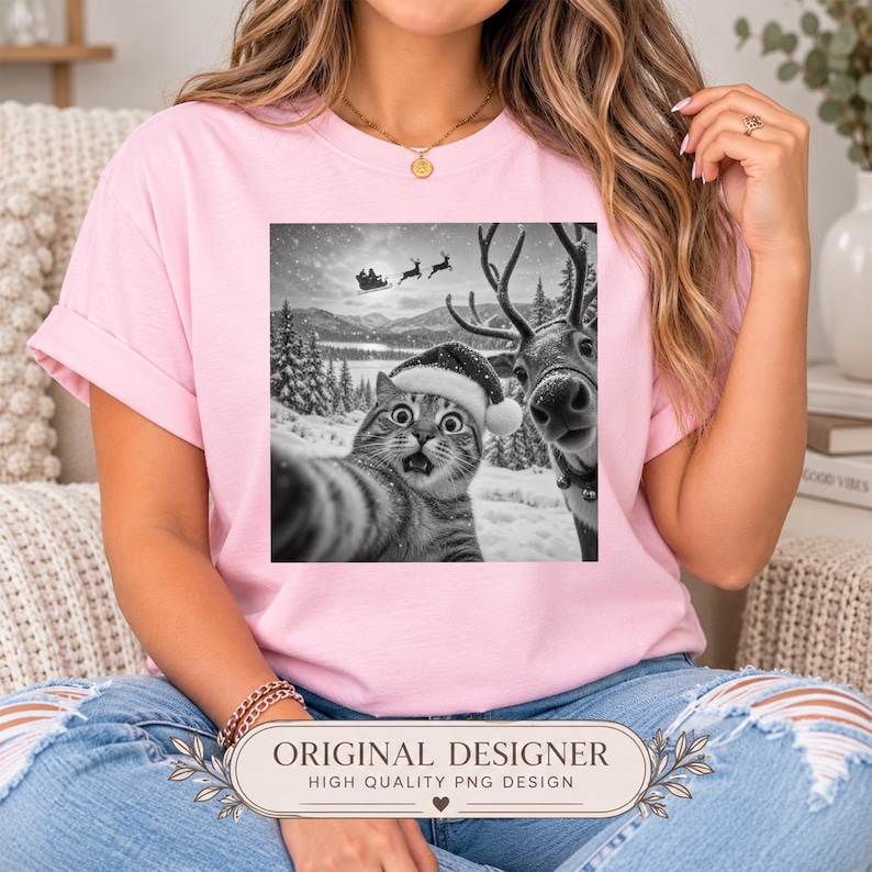 May include: A light pink short-sleeved t-shirt featuring a black and white graphic print of a cat wearing a Santa hat taking a selfie with a reindeer in a snowy landscape, with Santa's sleigh flying in the background sky above the trees.