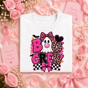 May include: A white t-shirt featuring a graphic of a ghost with a pink leopard print bow, the text BOO CREW, a black heart, and a checkered pattern. The ghost has a pink bubble, and the design includes small orange stars and black bats.