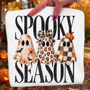 Può includere: Una t-shirt bianca con il testo nero SPOOKY SEASON e tre fantasmi. Il primo fantasma ha fiori arancioni e pipistrelli neri, il secondo una stampa leopardata con un fiocco a quadri e il terzo un motivo a scacchi bianco e nero con una zucca.